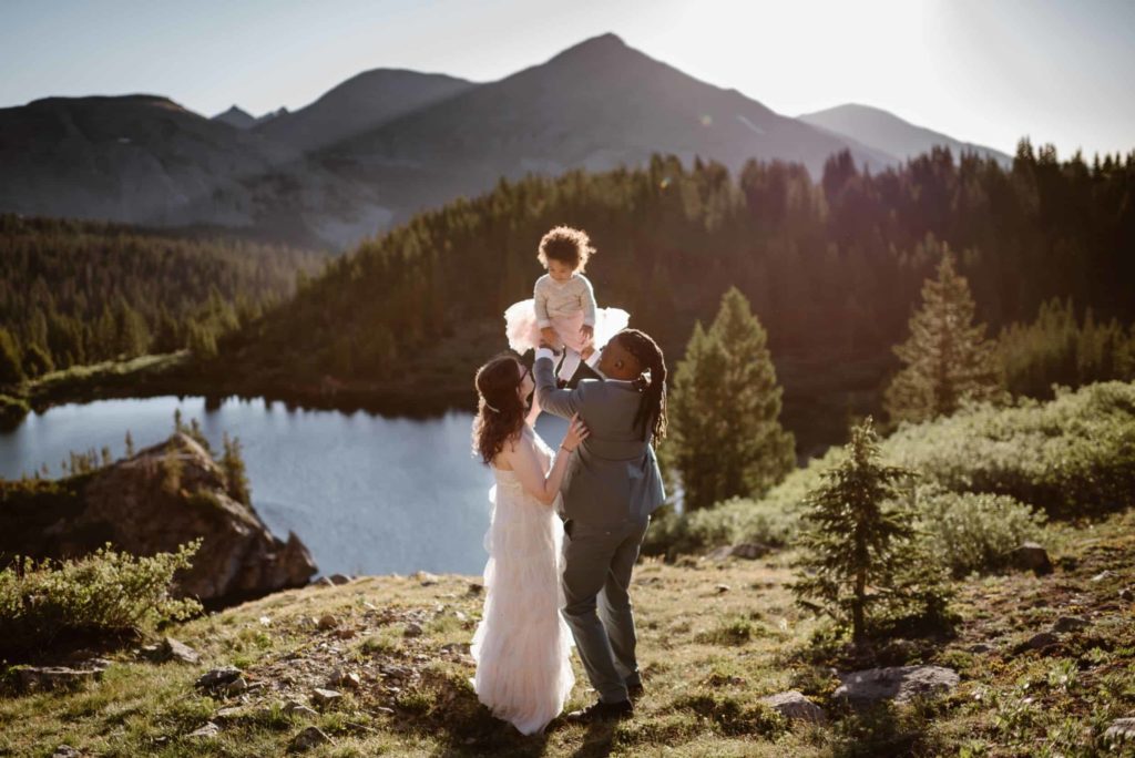 Bride and groom hold up their child, with lake, forest, and mountains in the background. 