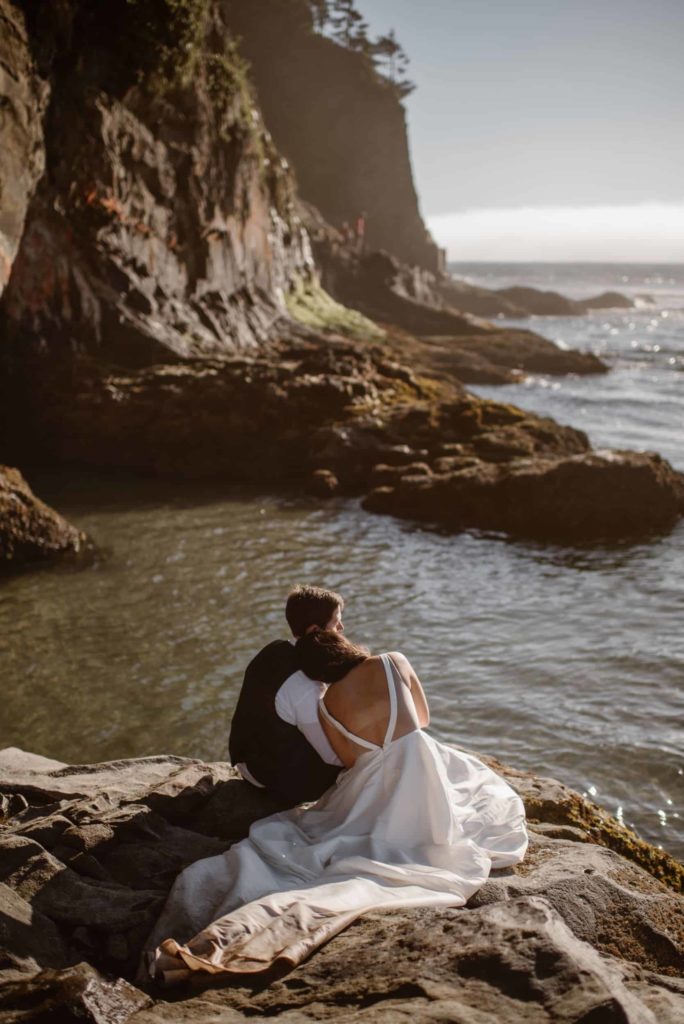 Bride and groom sit on rock and look out at the ocean. 