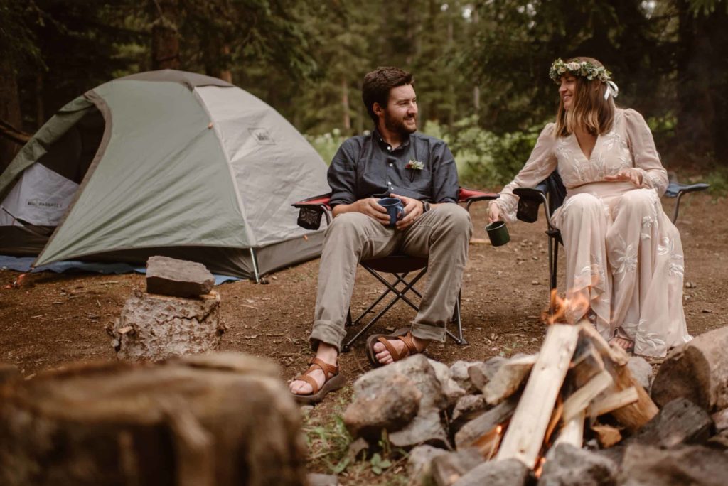 Bride and groom sitting in front of a campfire and smiling at each other. Their tent is set up behind them. 