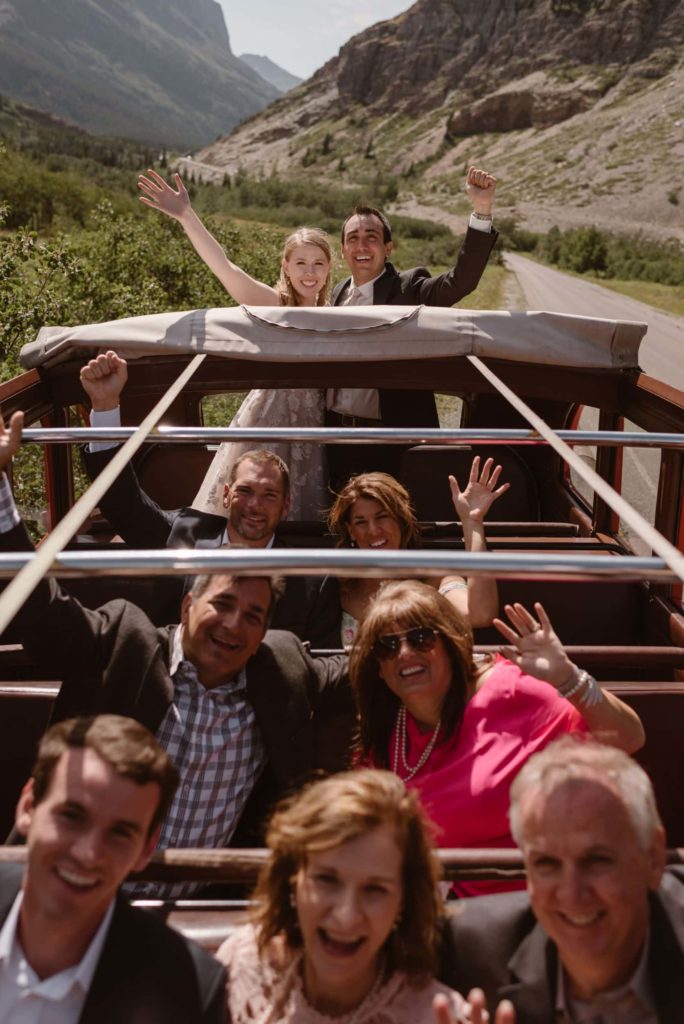 A bride and groom ride on the back of an automobile while their family and friends sit in the front while waving at the camera.