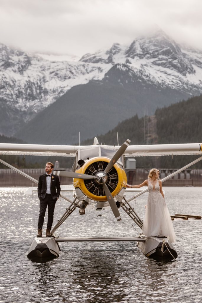 Bride and groom stand on outside of floatplane, while it rest on the water, with mountains in the background. 