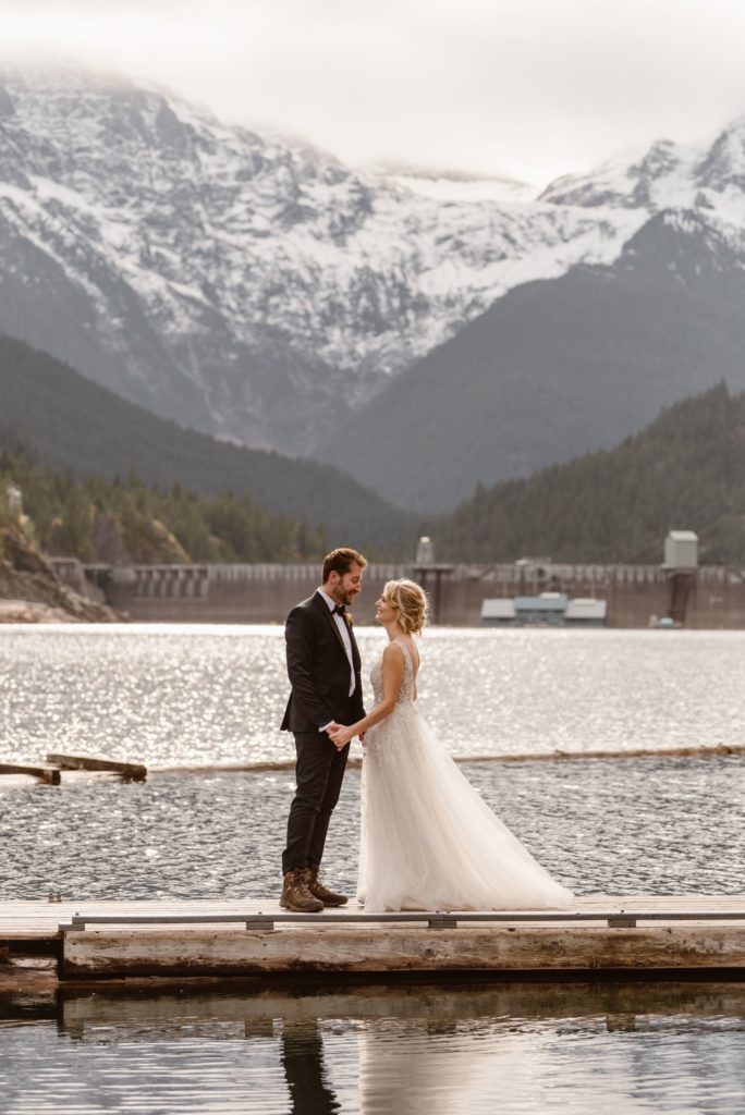 Bride and groom face each other while holding hands and standing on dock. 