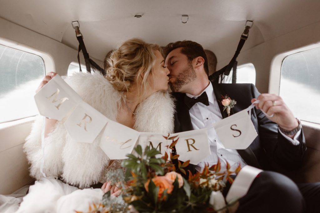 Bride and groom sit in floatplane, kissing, and hold up banner that says "Mr. and Mrs."