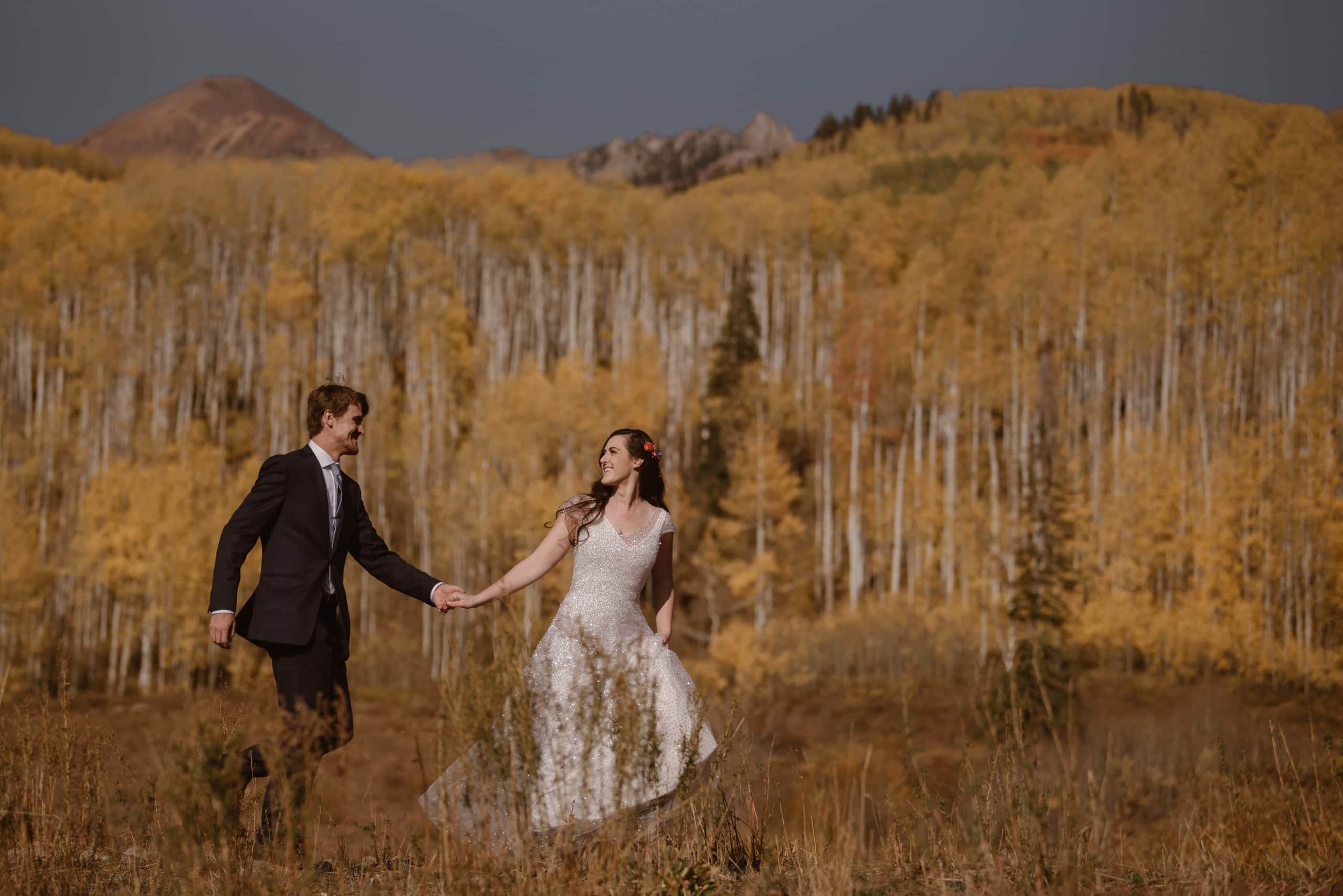 Bride and groom hold hands and walk through meadow in Crested Butte, Colorado. There is an aspen grove in the background with golden yellow leaves. 