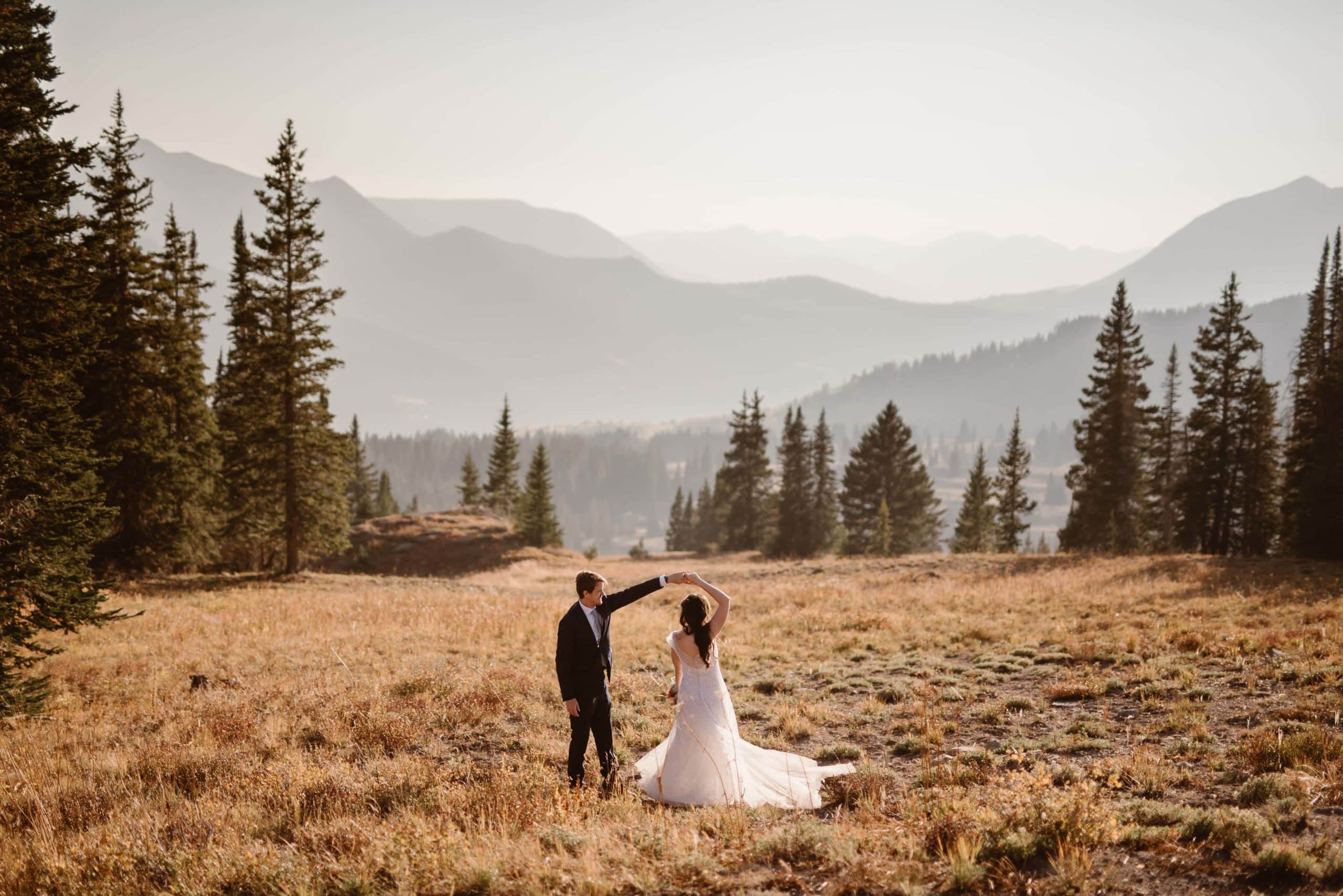 Groom twirls bride in a meadow in Crested Butte, Colorado. There are forests and mountains in the background.