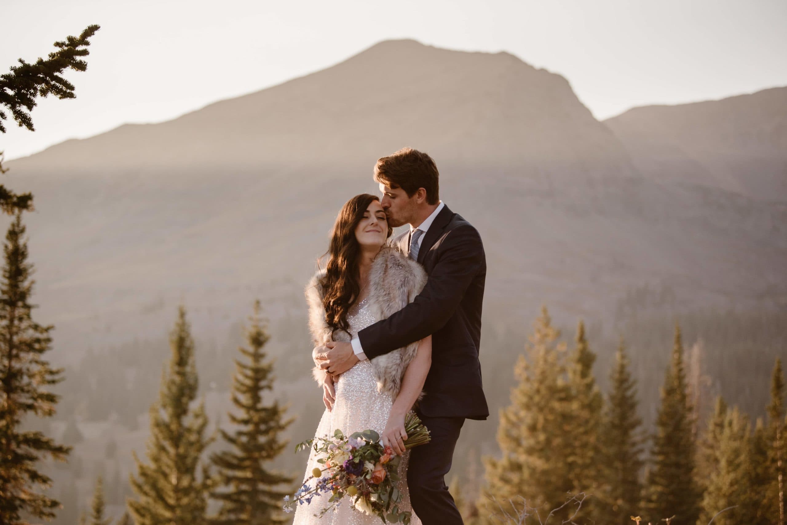 Bride and groom embrace on their elopement day in Crested Butte, Colorado. Groom kisses bride on the forehead. There are trees and mountains in the background. 